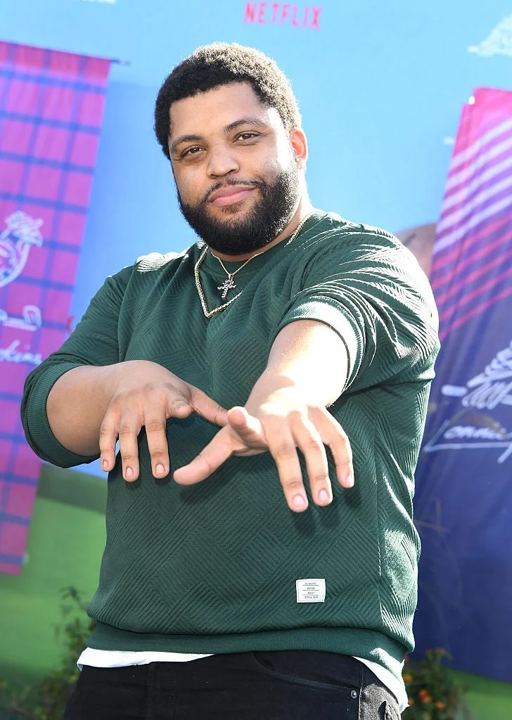 I don't know who this person is, but they are posing outdoors with a casual style, wearing a textured sweater and showing playful hand gestures
