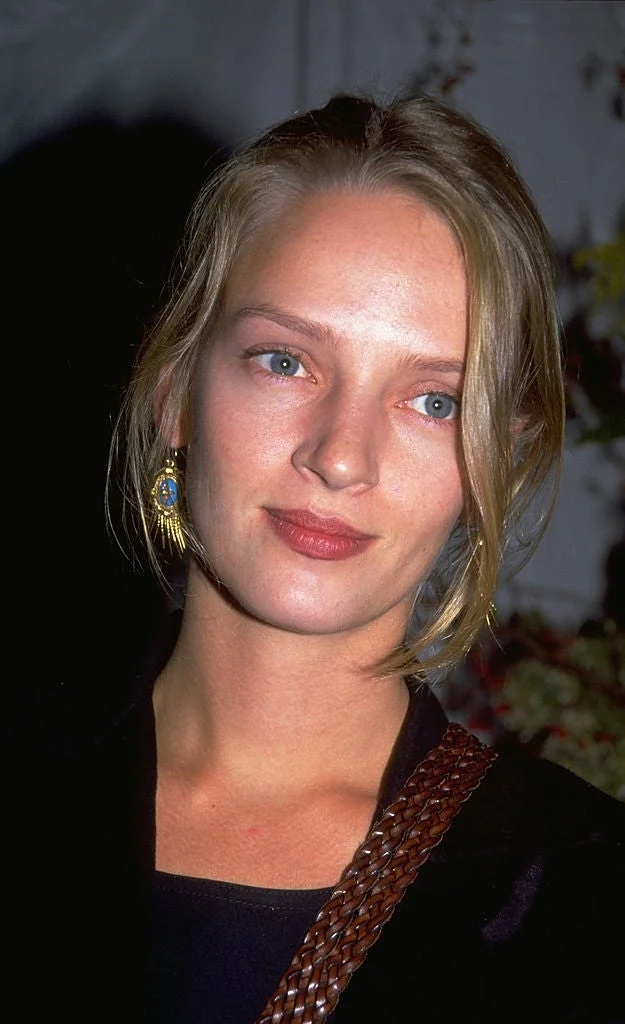 A person with light hair wearing a sleek black outfit, accessorized with statement earrings, appears in a celebrity setting