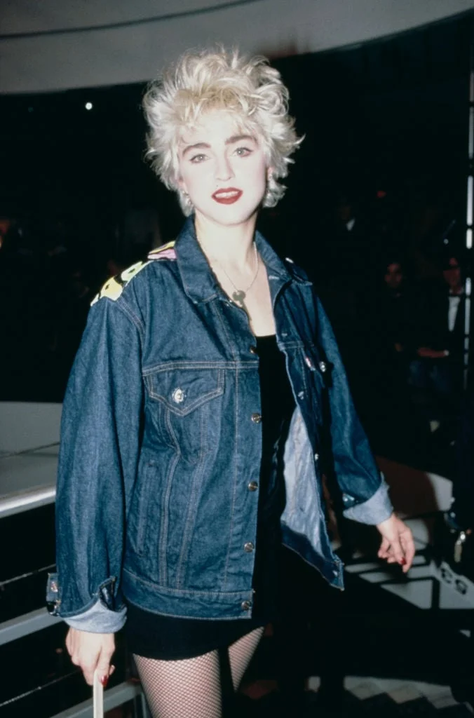 A person in a denim jacket with styled hair and bold makeup on a red carpet