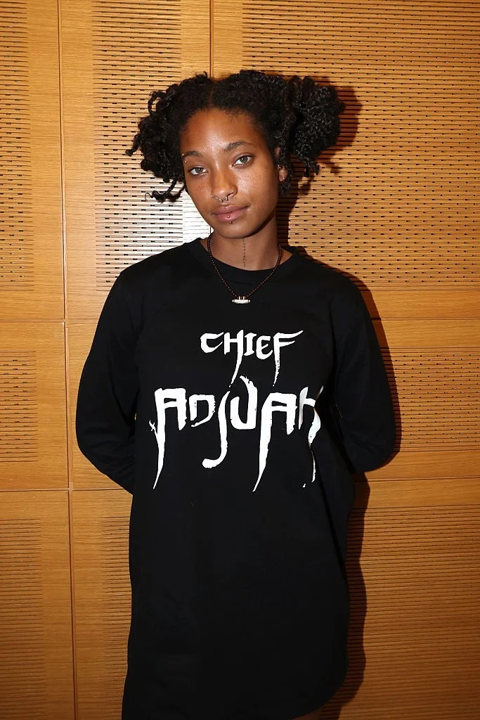 Person wearing a long-sleeve shirt with "Chief Adjuah" text, standing against a wooden backdrop, with braided hairstyle and nose ring