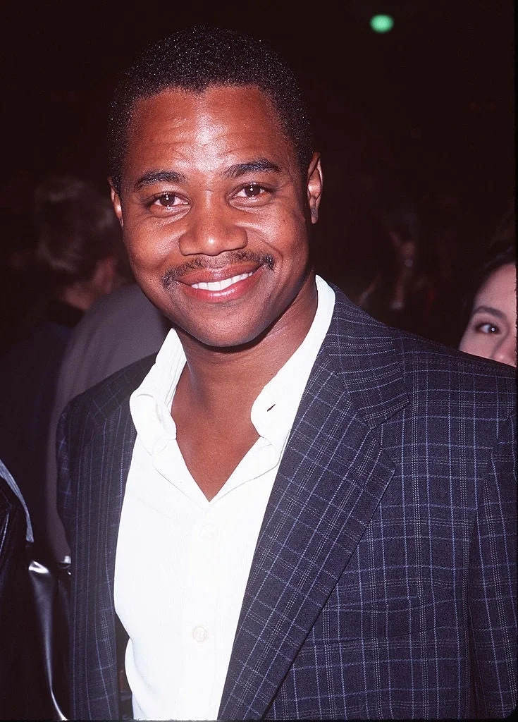 Man in a plaid suit jacket and open-collared white shirt smiles for the camera at a formal event