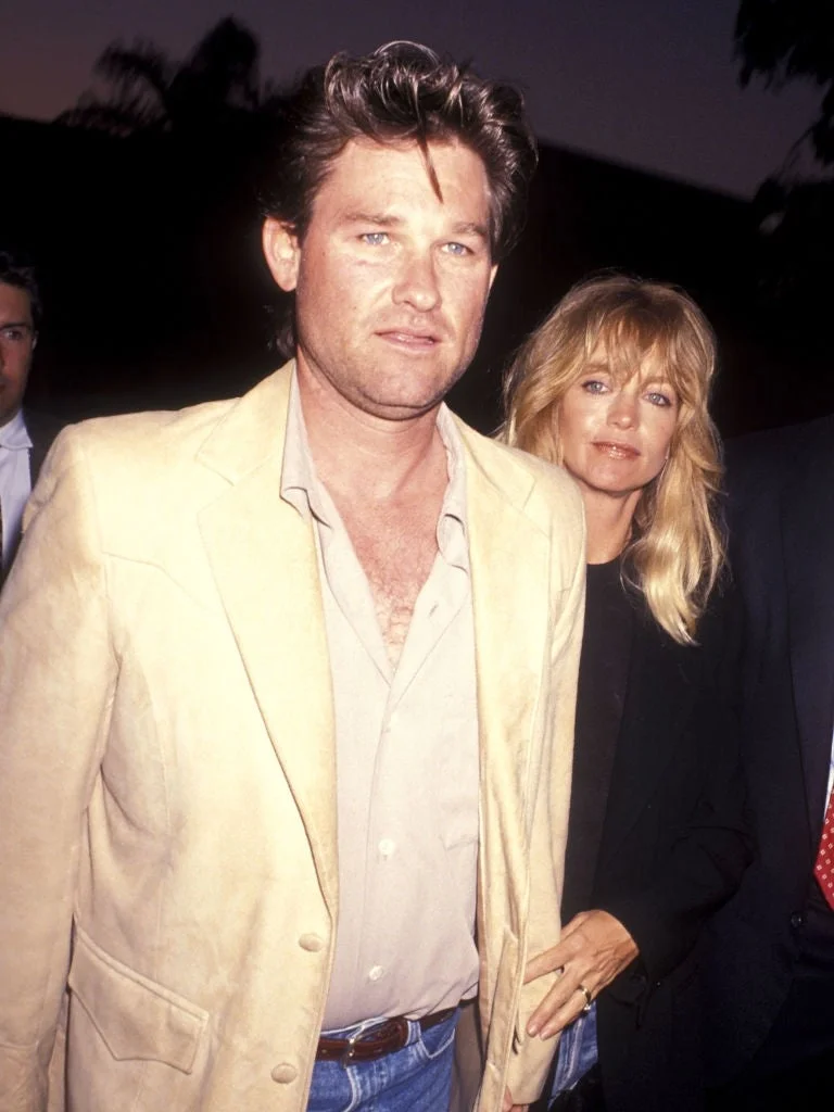 A man in a casual blazer and shirt stands with a woman in a black outfit. They are at a public event, possibly a red carpet appearance
