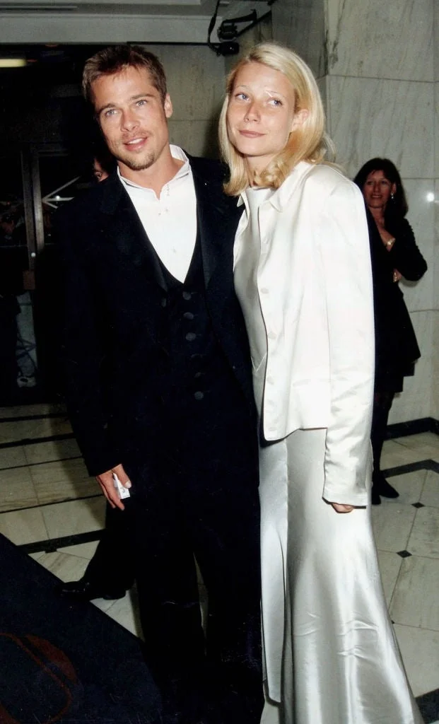 Two people on a red carpet; one in a black suit and the other in a light satin outfit with a jacket