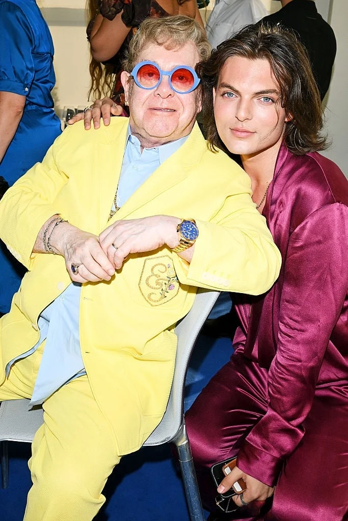 Two individuals seated together. The person on the left wears a bright suit and round glasses. The person on the right wears a silky suit