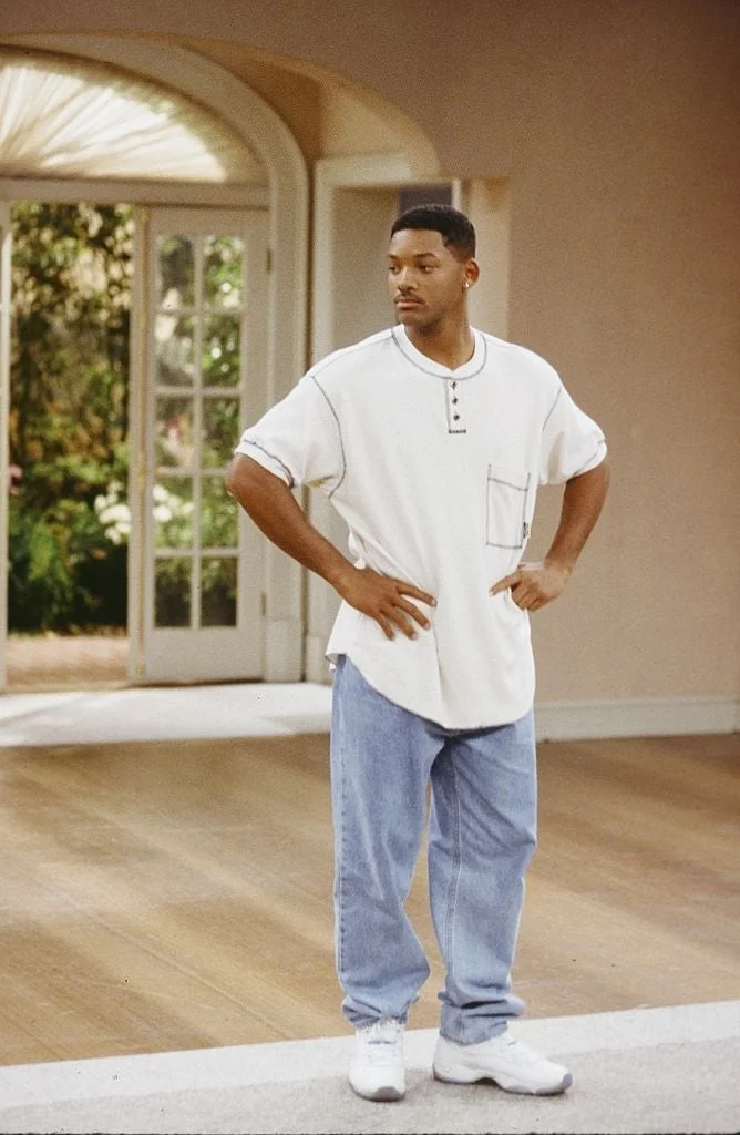 Person standing casually indoors, wearing a loose white shirt, light blue jeans, and sneakers, with hands on hips