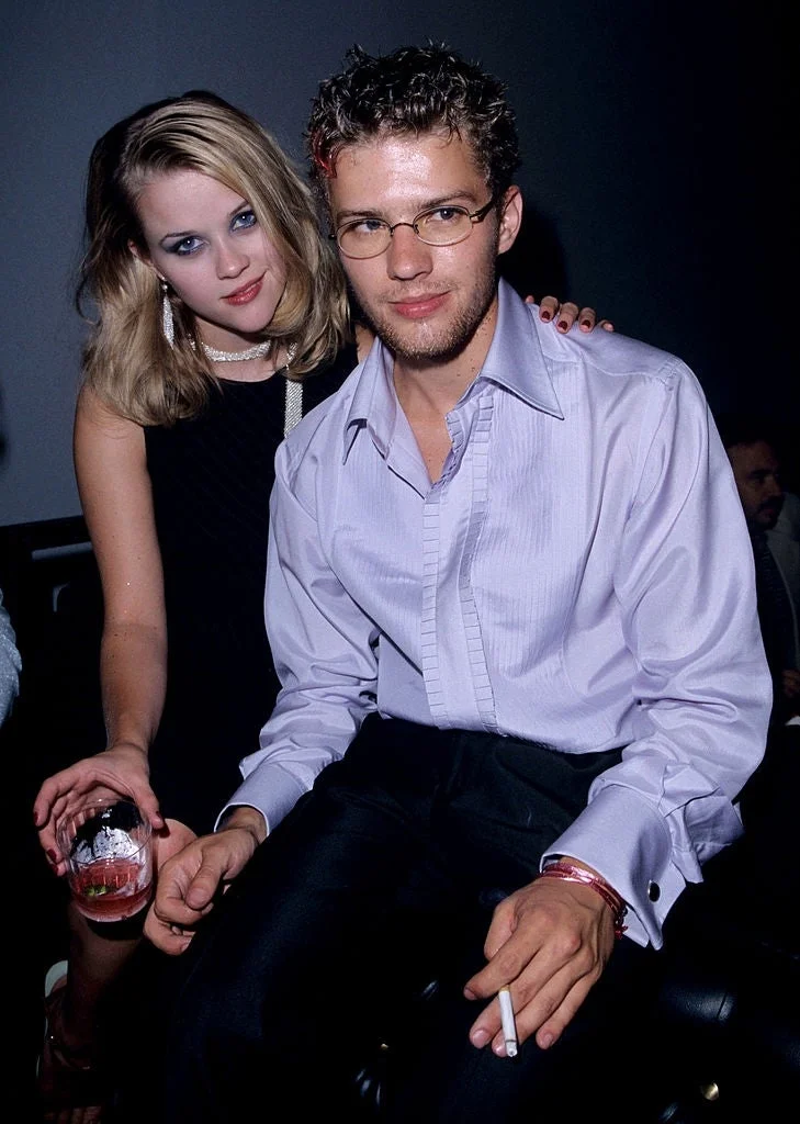 Two celebrities at an event; the woman wears a chic sleeveless dress, and the man in a smart-casual button-up shirt holds a cigarette