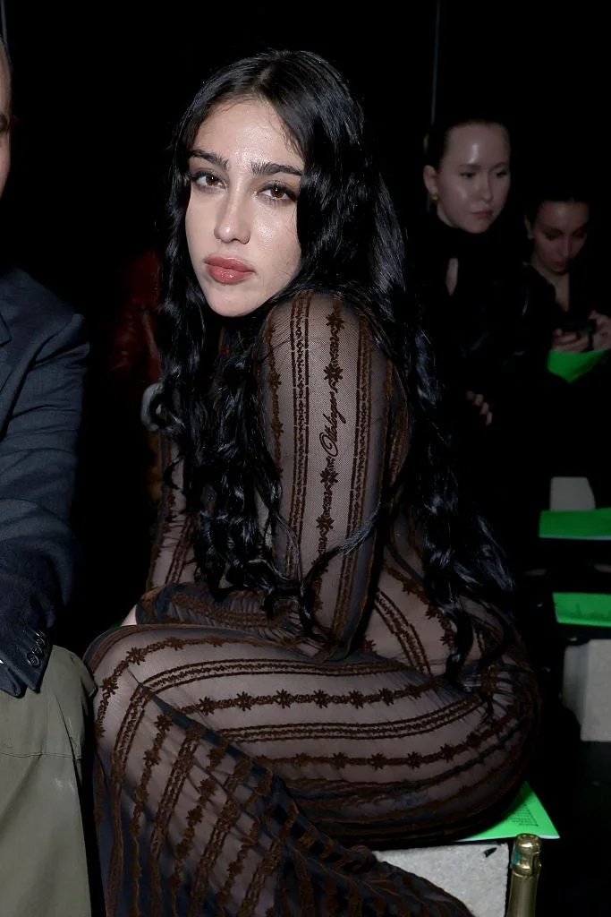 Person seated wearing a sheer, patterned outfit with long hair, looking at the camera