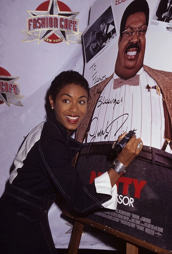 A person in a stylish suit poses with a poster for "The Nutty Professor," signing it with a marker at a promotional event