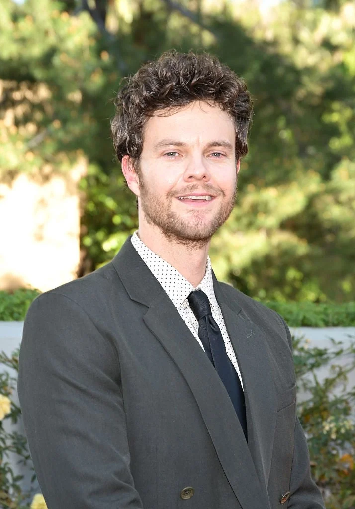 A person in a suit with a polka dot shirt stands outdoors at an event, smiling