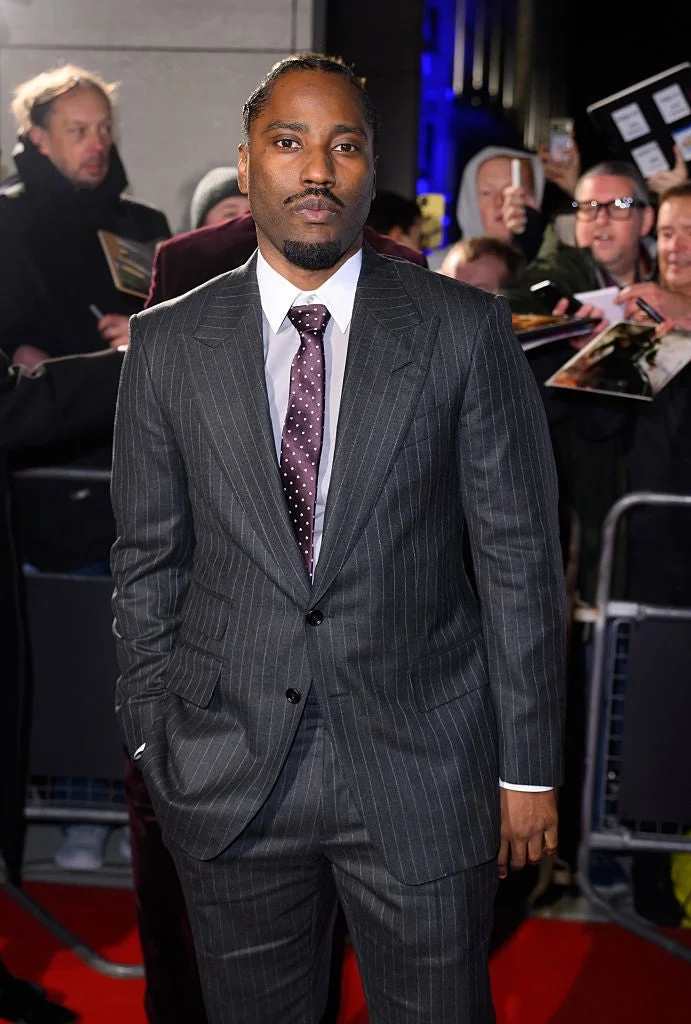 Person in a pinstripe suit and polka dot tie on a red carpet, with fans in the background holding items for signing