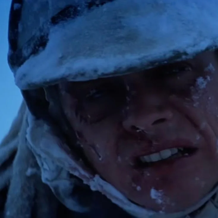 A person wearing winter gear is covered in snow, appearing determined and focused, possibly in a snowy wilderness setting