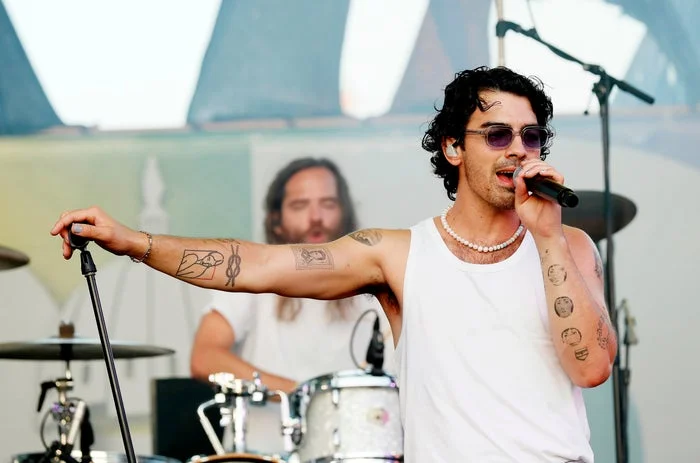 Person singing on stage, wearing sunglasses, a white tank top, and a beaded necklace, with another band member playing drums in the background