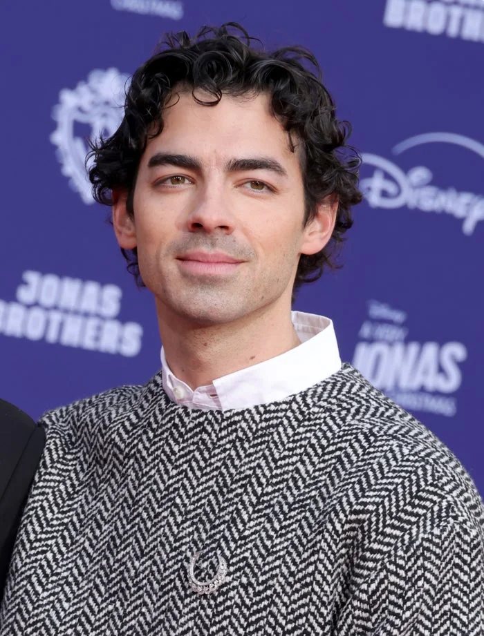 Person with curly hair wearing a textured sweater over a collared shirt, standing in front of a backdrop with logos