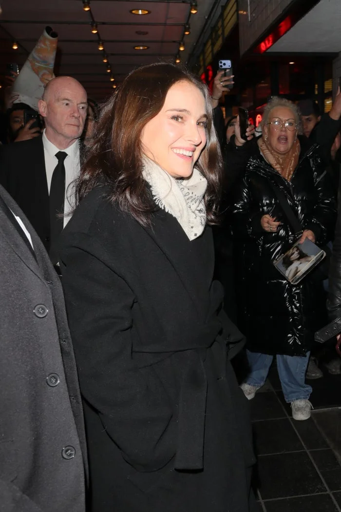 A celebrity in a stylish black coat smiles at the crowd while walking, surrounded by fans and security