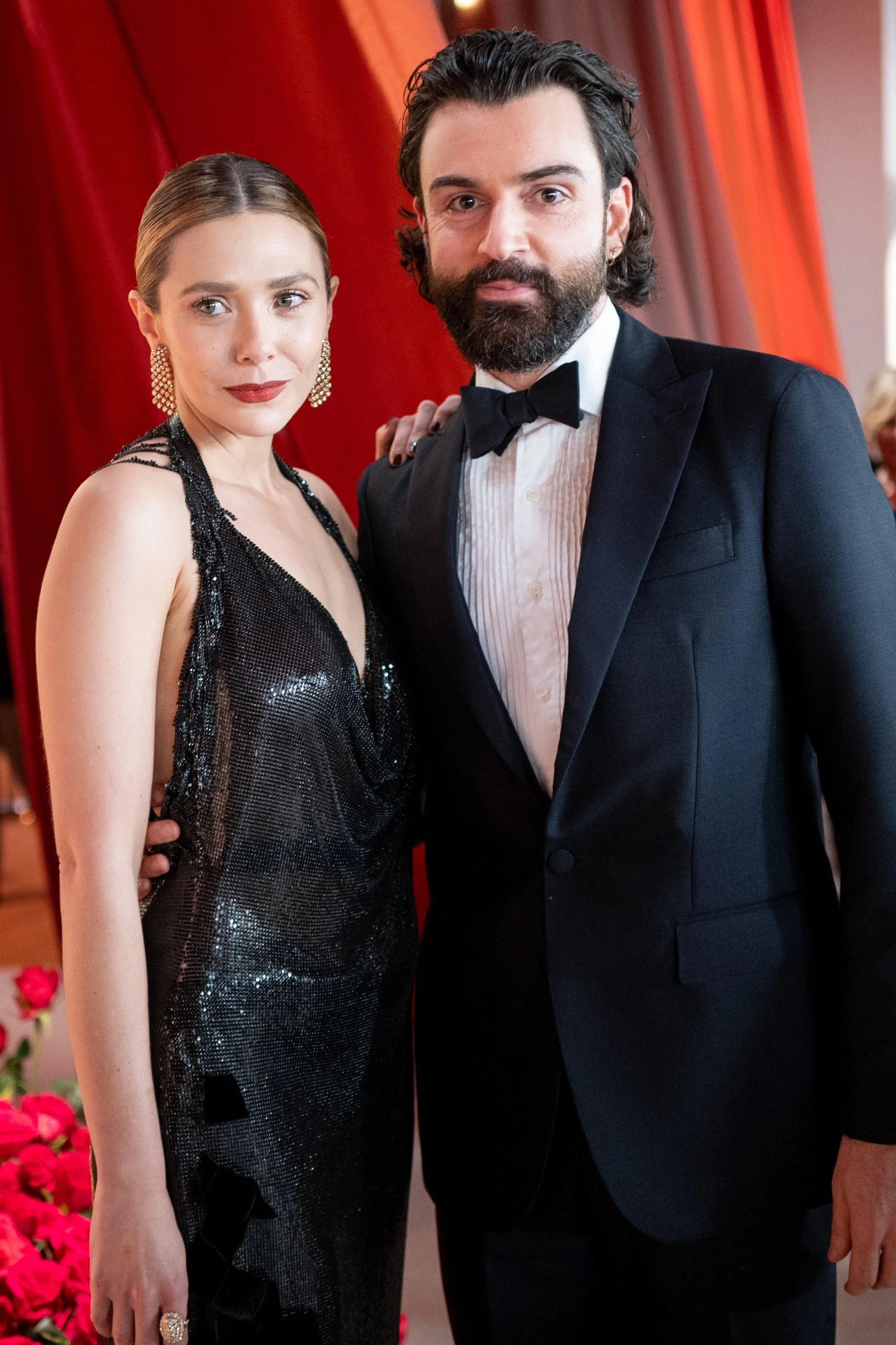 Two individuals pose at a formal event; the woman wears a sequin gown, and the man is in a classic tuxedo