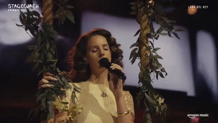 Lana Del Rey performing on stage with floral decor, holding a microphone.