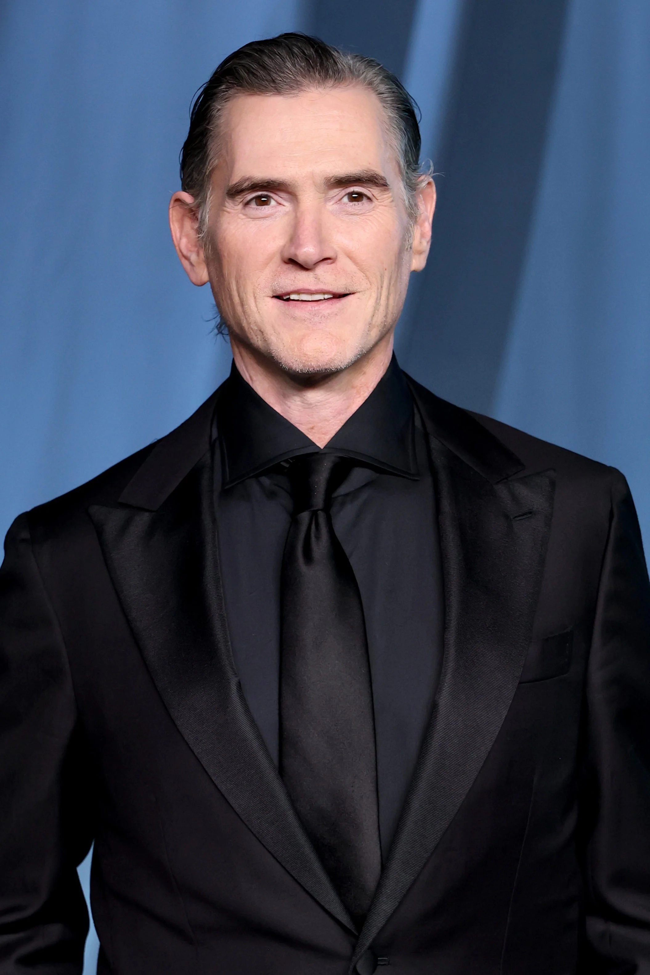 Person in a tailored black suit and tie stands on a formal event backdrop, hands in pockets