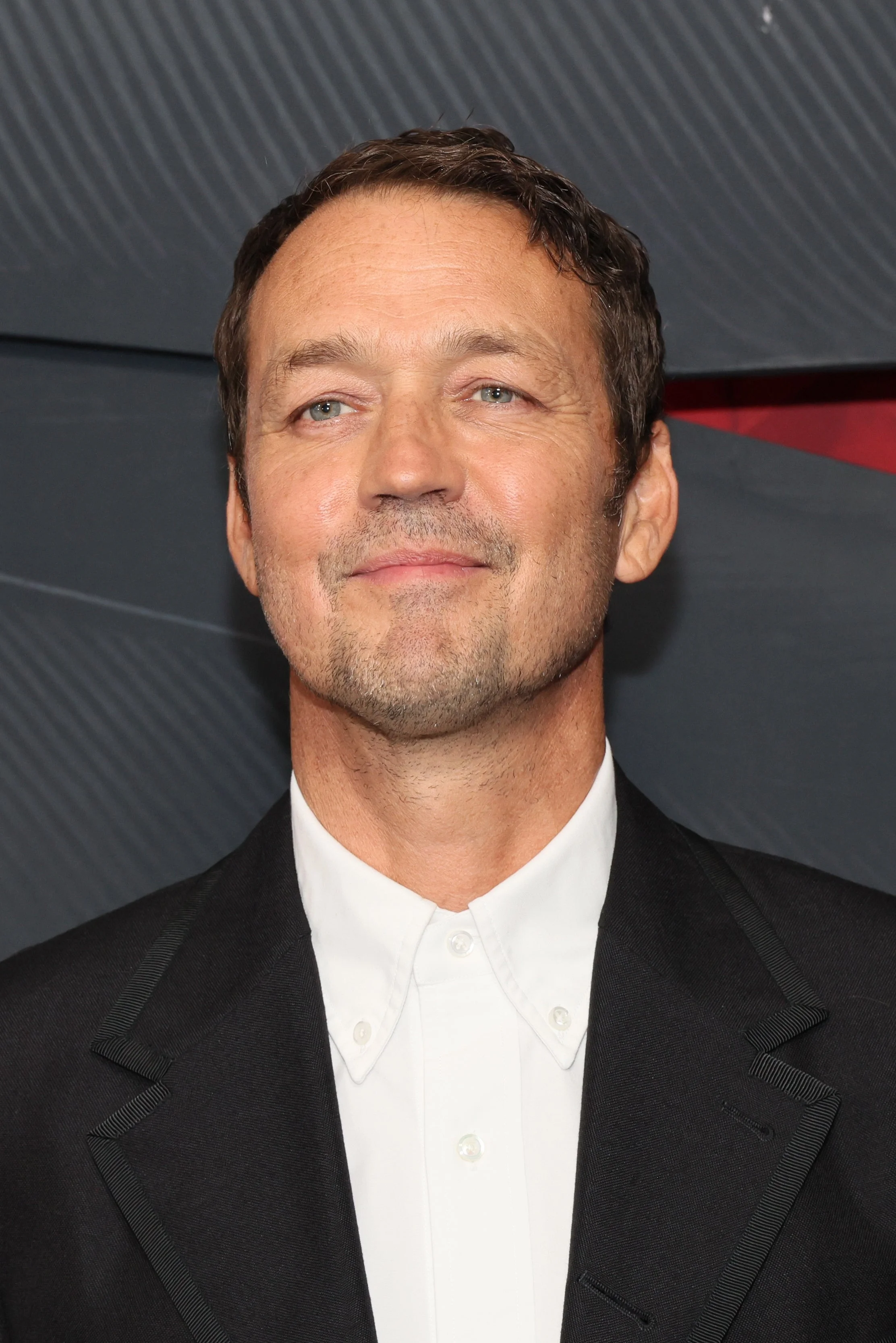 Person smiling on a red carpet, wearing a classic black suit with a white shirt