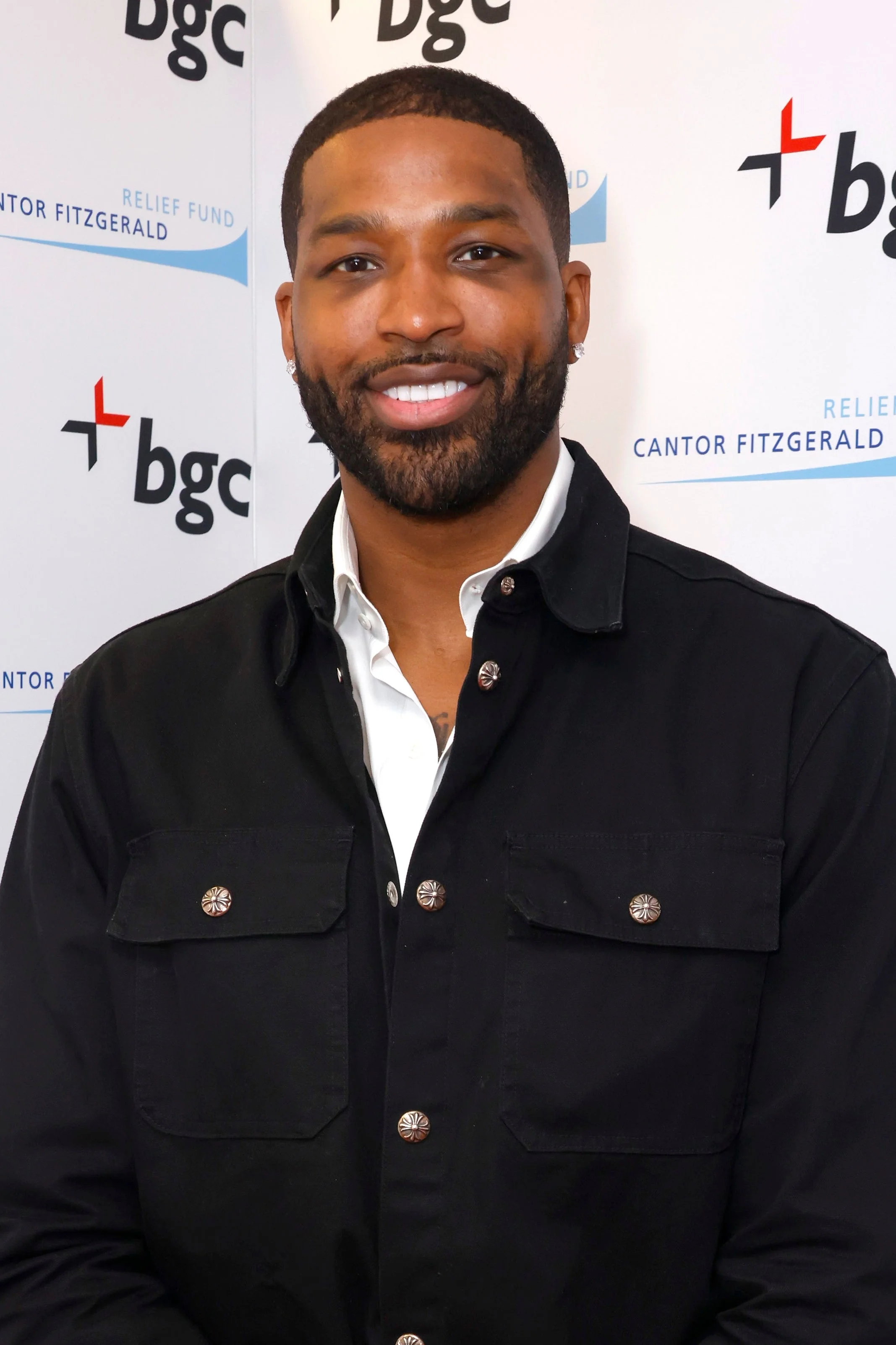 A man smiles in a casual button-up jacket and shirt at a charity event backdrop with logos