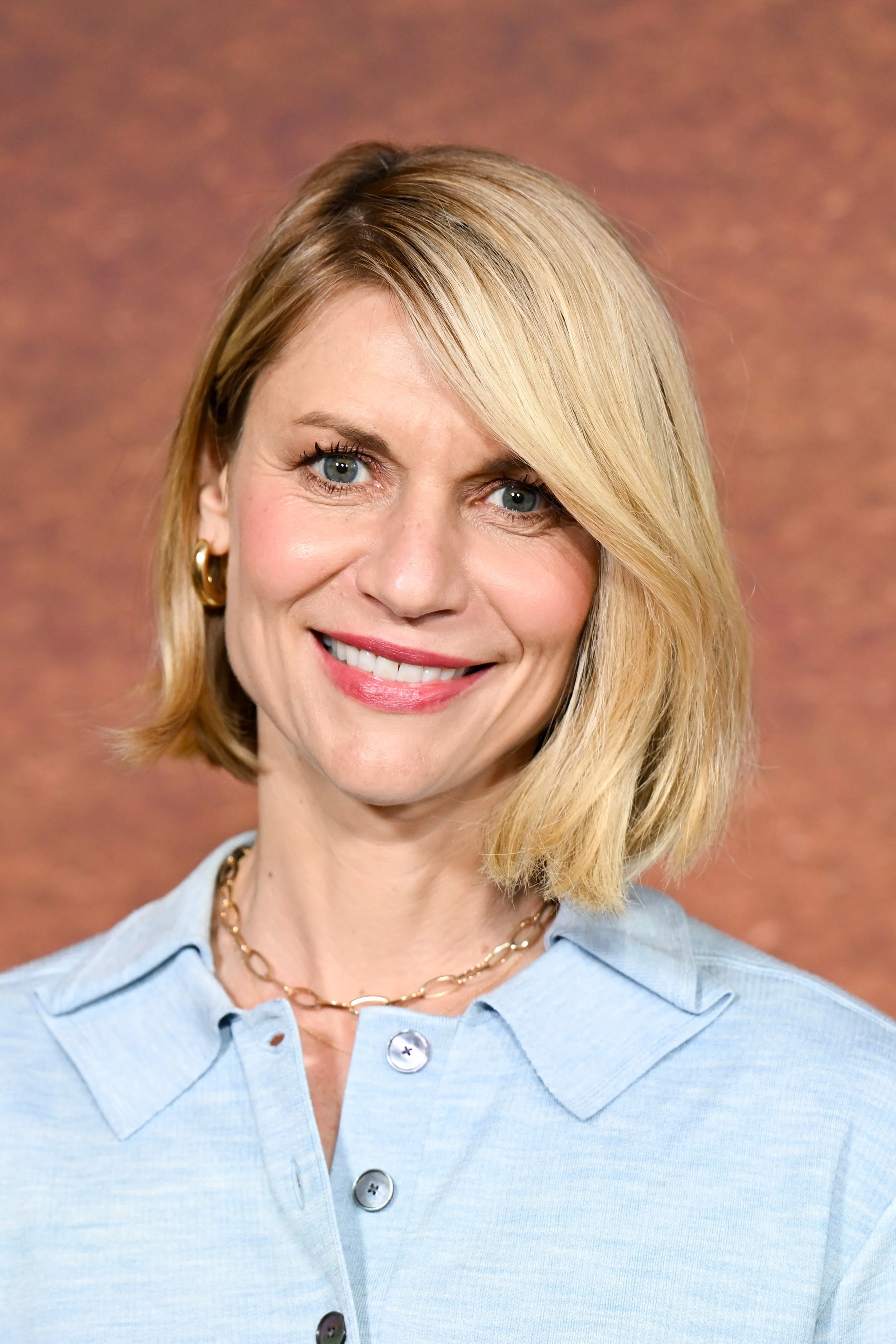 Person with short, sleek hairstyle wears a button-up shirt, smiling in front of a neutral background
