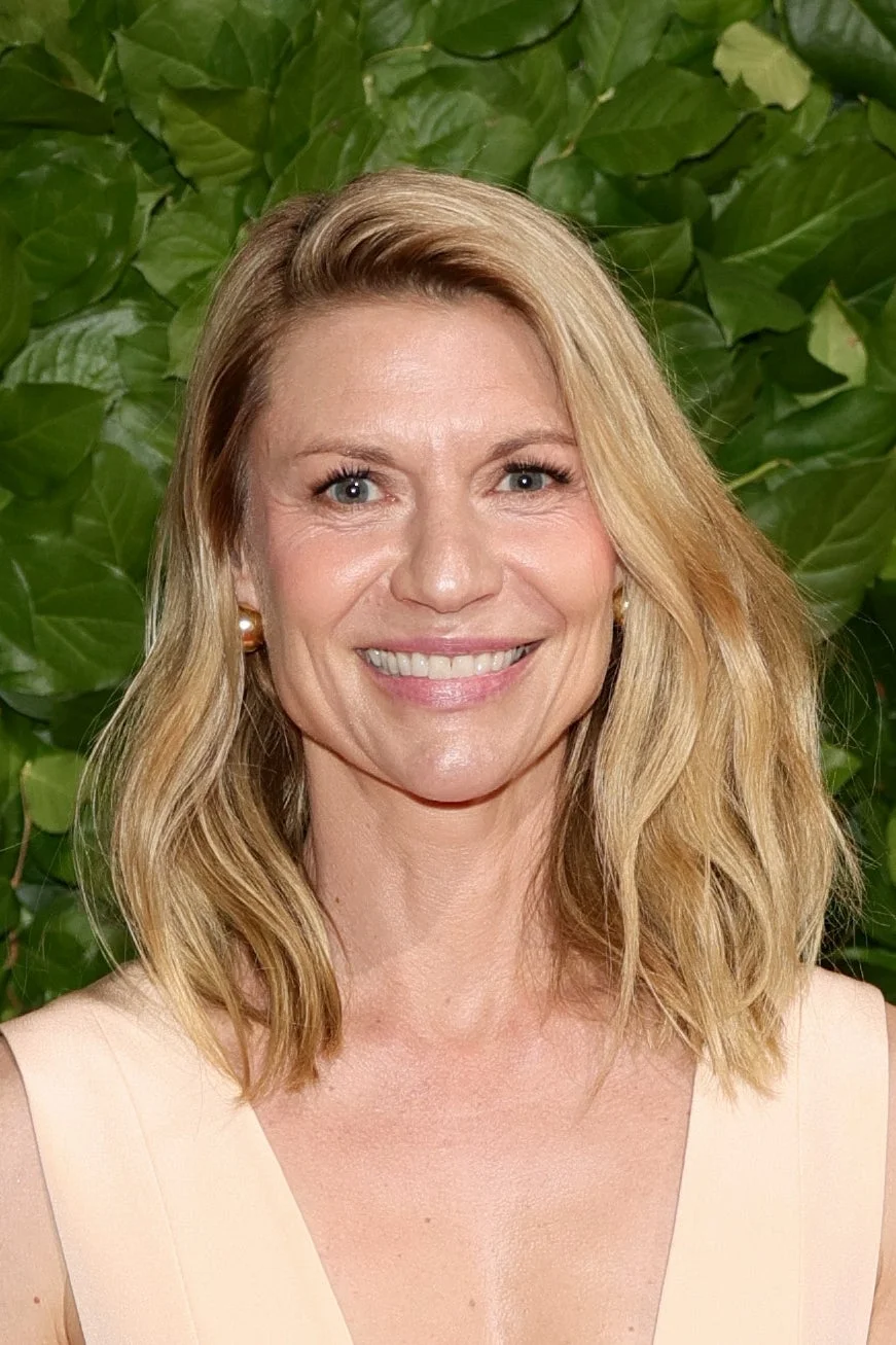 Celebrity on a green leafy background, wearing a sleeveless, elegant dress and smiling at the camera