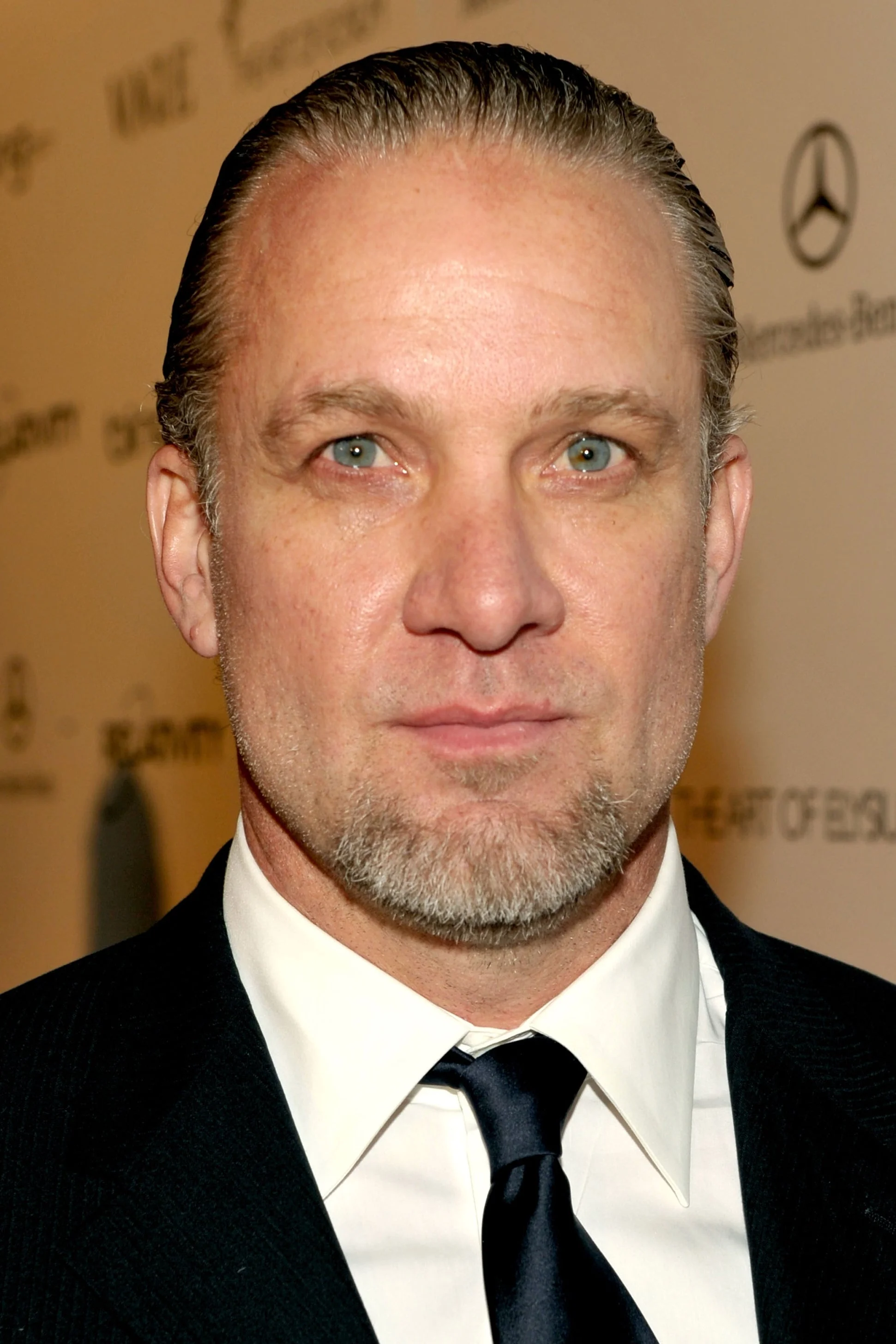 Celebrity in a suit and tie poses for a photo at an event, standing against a backdrop with logos