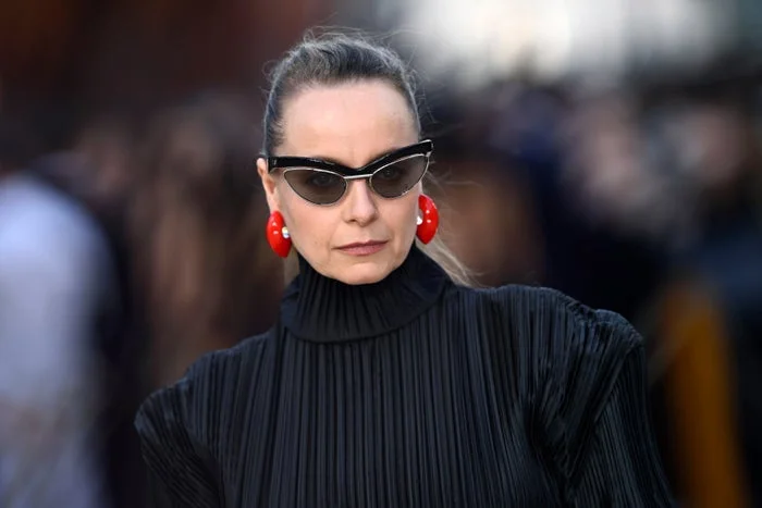 A person wearing a high-neck pleated black outfit, cat-eye sunglasses, and bold red earrings at a celebrity event