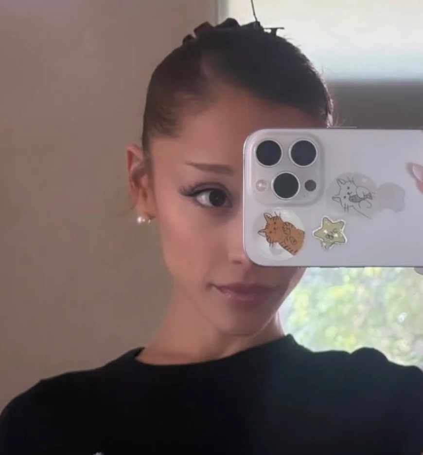 Person taking a mirror selfie, partially obscured by a phone with cat stickers. They have styled hair and pearl earrings