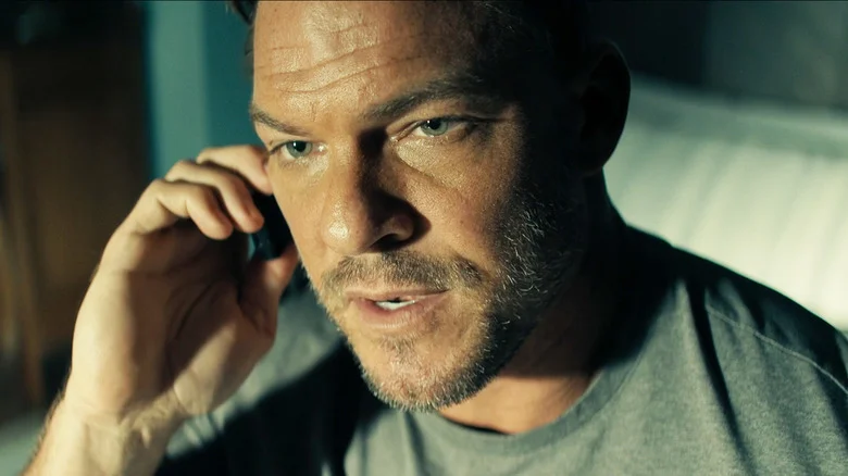 Alan Ritchson's Jack Reacher looks serious in closeup as he talks on a cell phone in Reacher