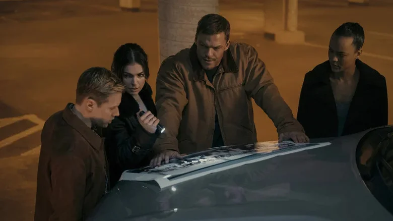 Shaun Sipos' David O'Donnell, Serinda Swan's Karla Dixon, Alan Ritchson's Jack Reacher, and Maria Sten's Frances Neagley stand together while looking at a document in Reacher.webp