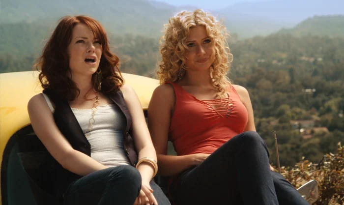 Two women sit on a car hood, casually dressed, with one speaking while the other looks on. They appear relaxed in an outdoor setting