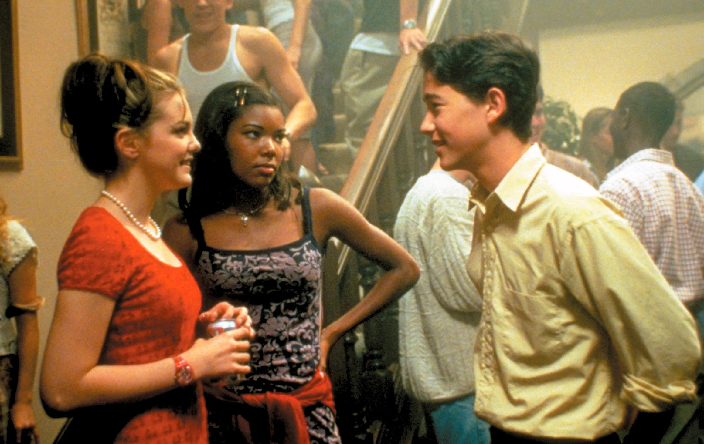 Three people converse at a lively house party on a staircase, surrounded by other guests