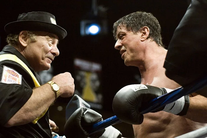 Two men in a boxing ring, one shirtless with gloves, and the other in a hat, talking intensely