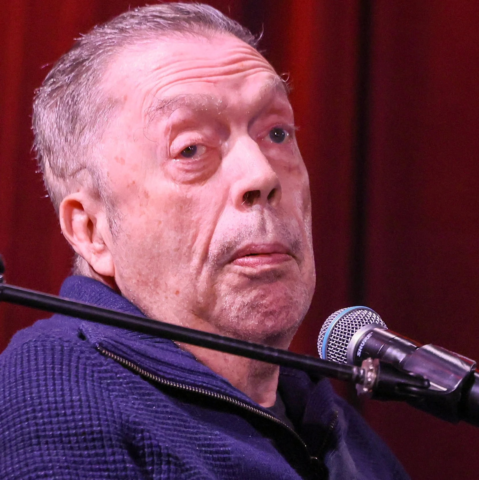 Tim Curry speaks into a microphone, seated against a red curtain backdrop, wearing a textured sweater