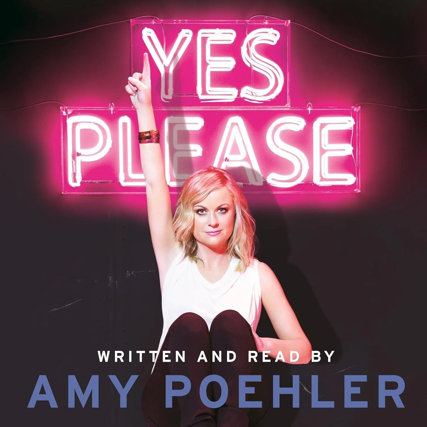 Amy Poehler sits under a neon "Yes Please" sign, raising her finger. She wears a white sleeveless top and dark pants