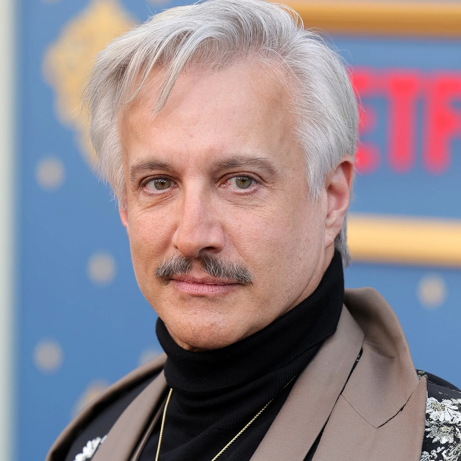 Bronson Pinchot in floral suit with a turtleneck and layered coat poses at a Netflix event