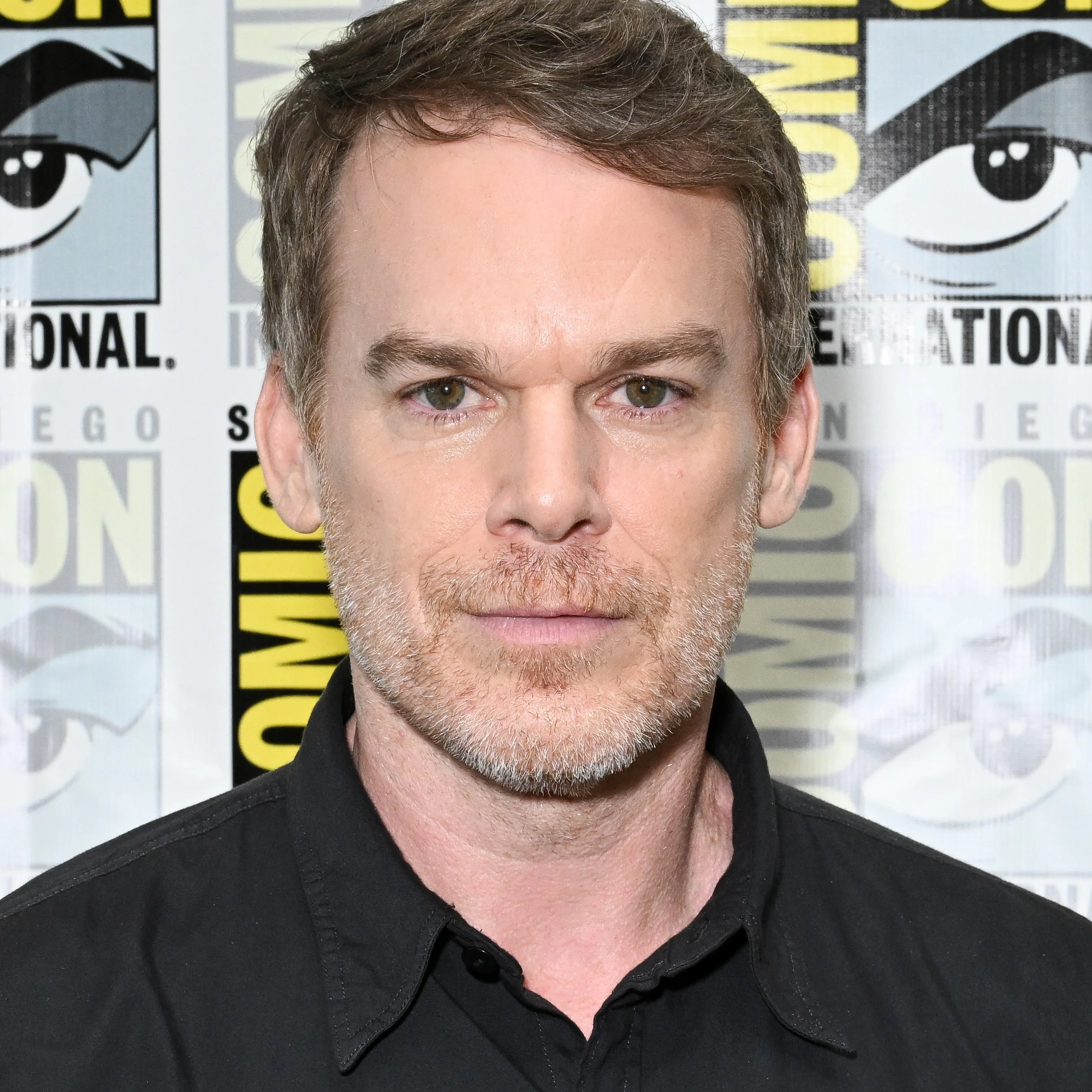 Michael C Hall in a black shirt at Comic Con, standing in front of backdrop with the event's logo