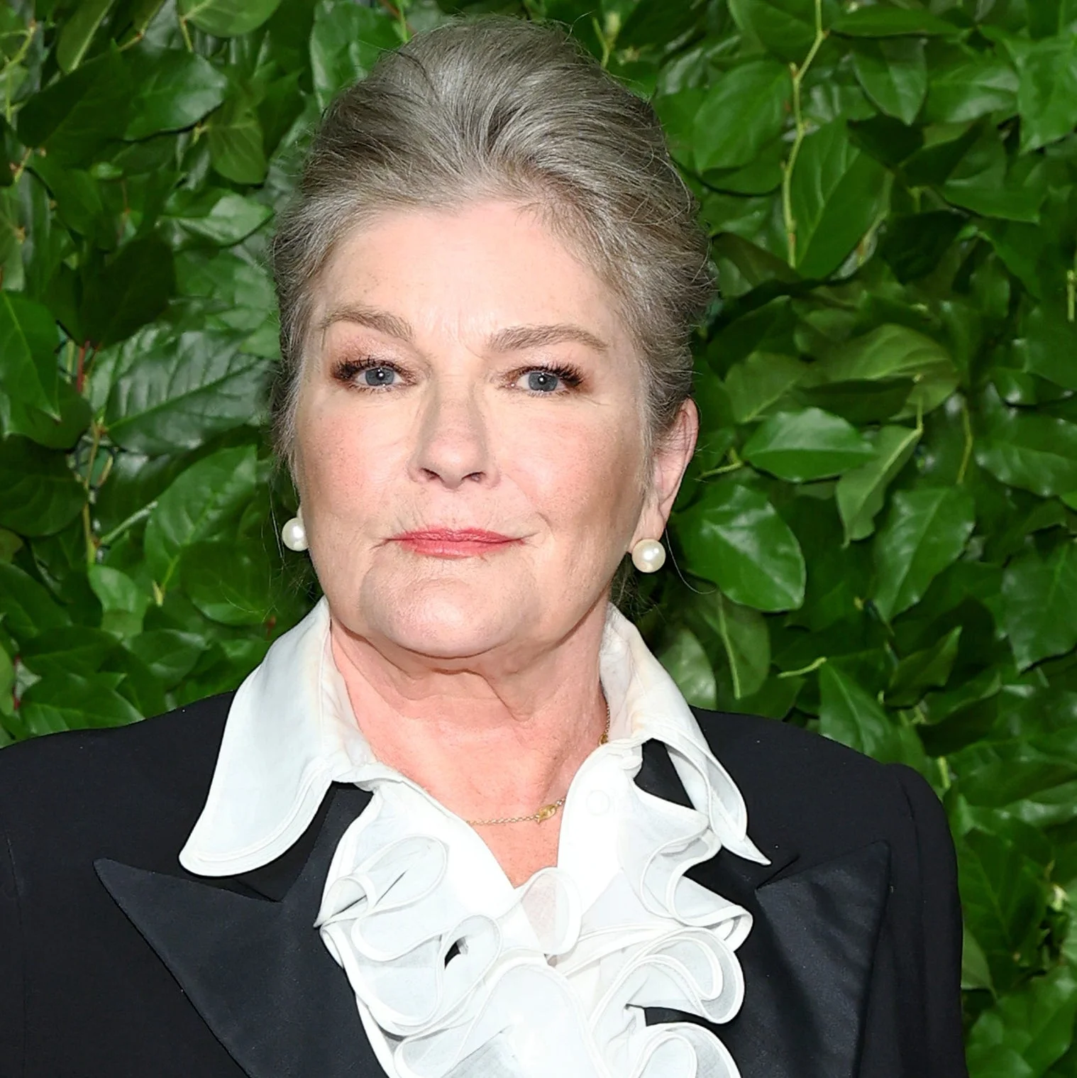 Person in a tailored suit with a ruffled shirt stands in front of a foliage wall at a formal event