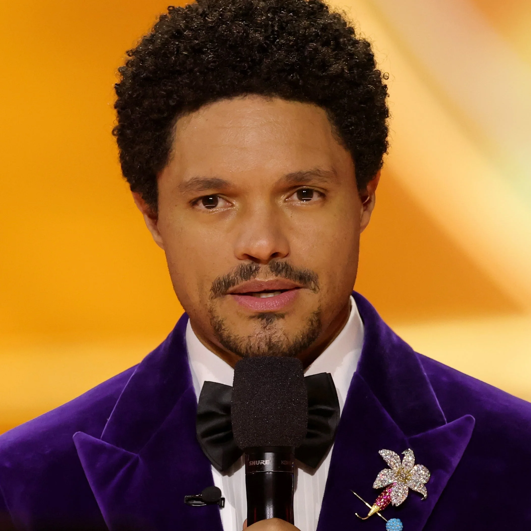 Trevor Noah in a velvet tuxedo holds a microphone on stage during an event