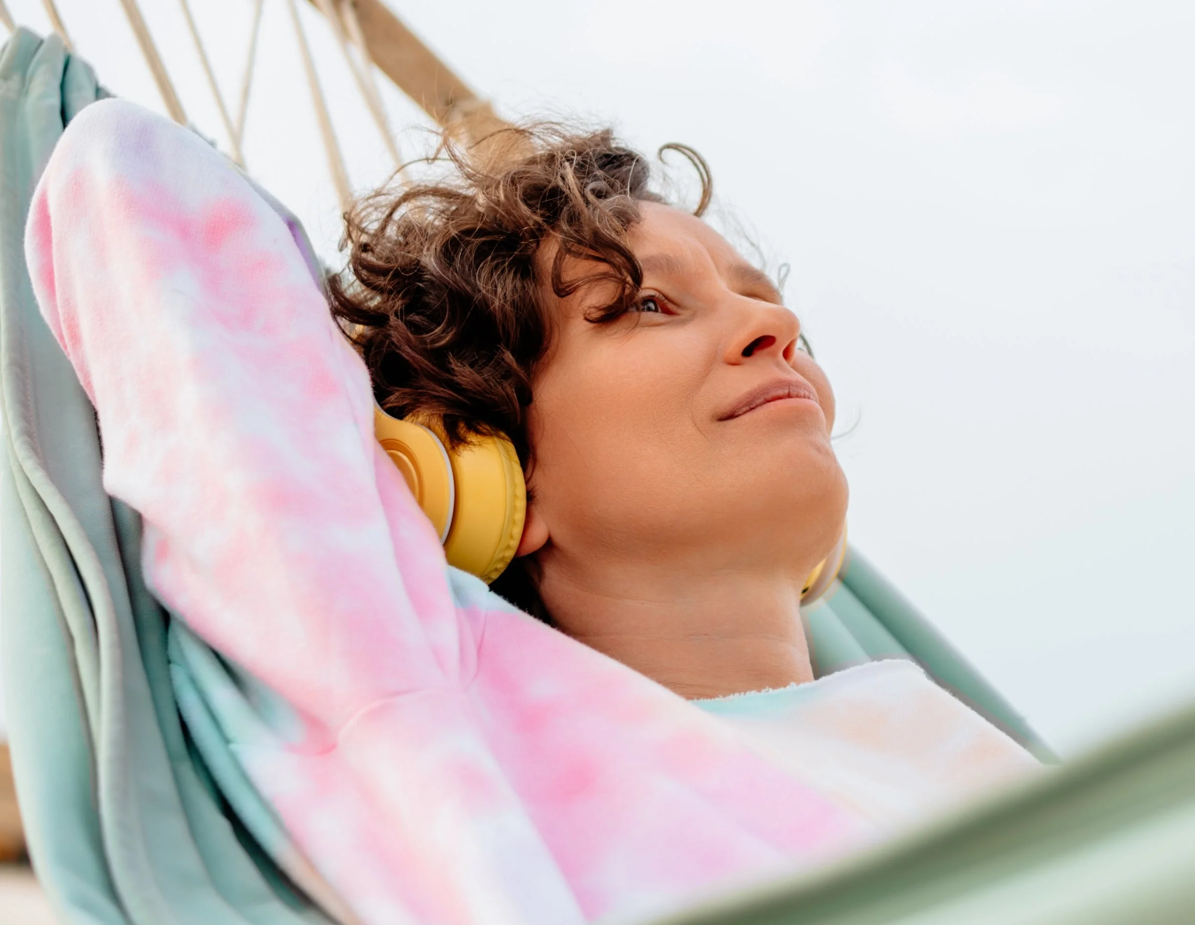 Person relaxing in a hammock under palm trees, wearing headphones and a tie-dye sweatshirt, looks content and peaceful