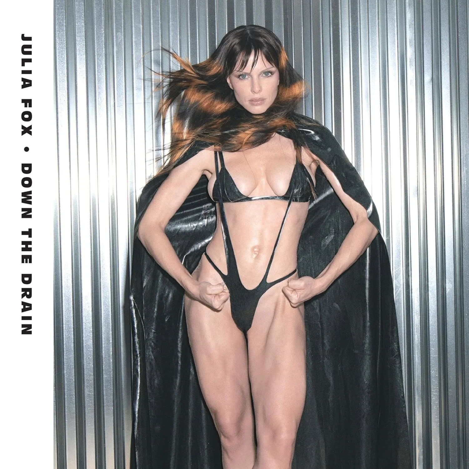 A person is posing confidently, wearing a strappy black outfit and cape, against a metallic background. The text reads "Julia Fox, Down the Drain."