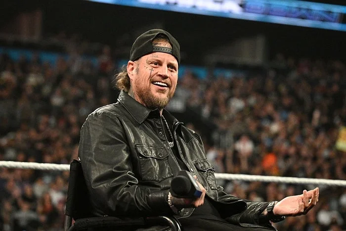 A person wearing a leather jacket and cap sits in a stadium, holding a microphone with a crowd in the background