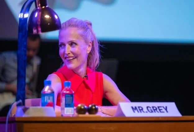 A person sits at a desk labeled "Mr. Grey" during an event, smiling and wearing a sleeveless top