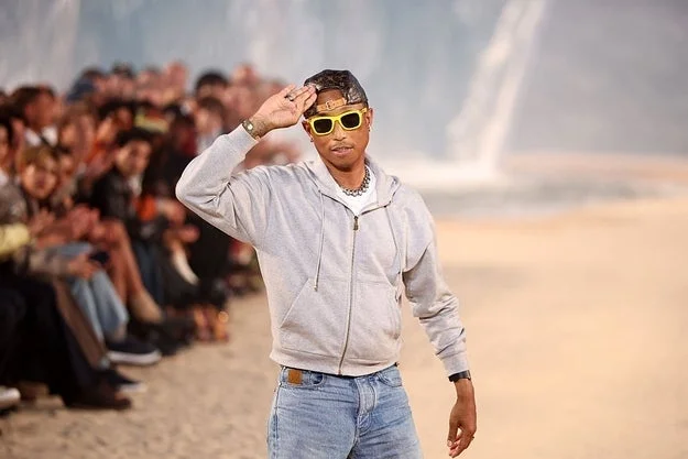 Person wearing casual attire, hoodie, jeans, and sunglasses, walking on a runway with a crowd in the background