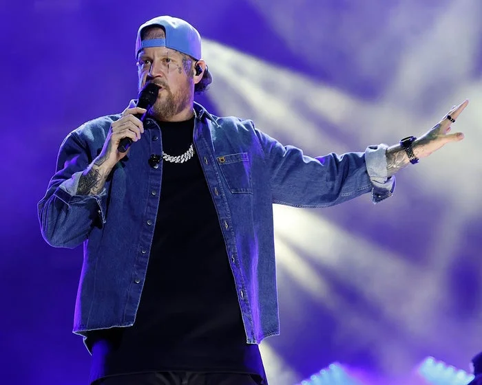 Person on stage in denim jacket and baseball cap, holding a microphone, gesturing with one hand during a performance