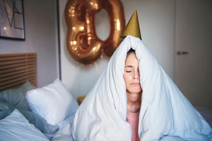 Person in bed with a duvet over their head, wearing a party hat. There's a large balloon shaped like the number 30 in the background