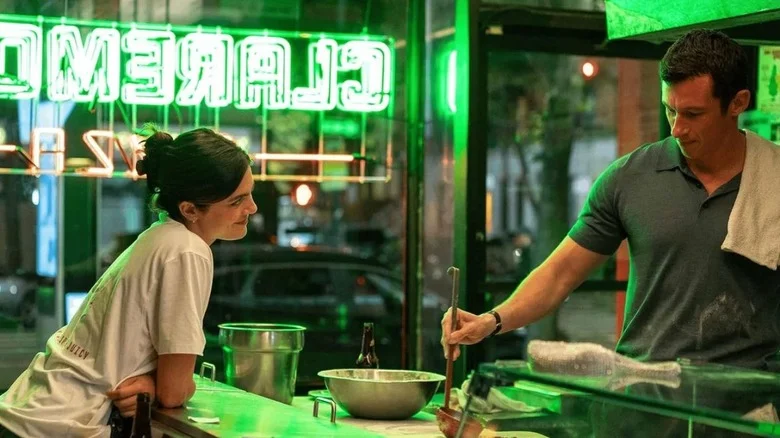 Monica Barbaro as Allie standing at the counter talking to Callum Turner as Owen as he makes a pizza in One Night Only