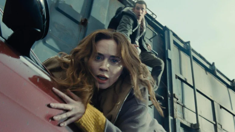 Emily Blunt as Margaret Fairchild in Disclosure Day, riding along the hood of a car next to a train while Josh O'Connor as Daniel Kellner hangs on to the train car ladder above her.