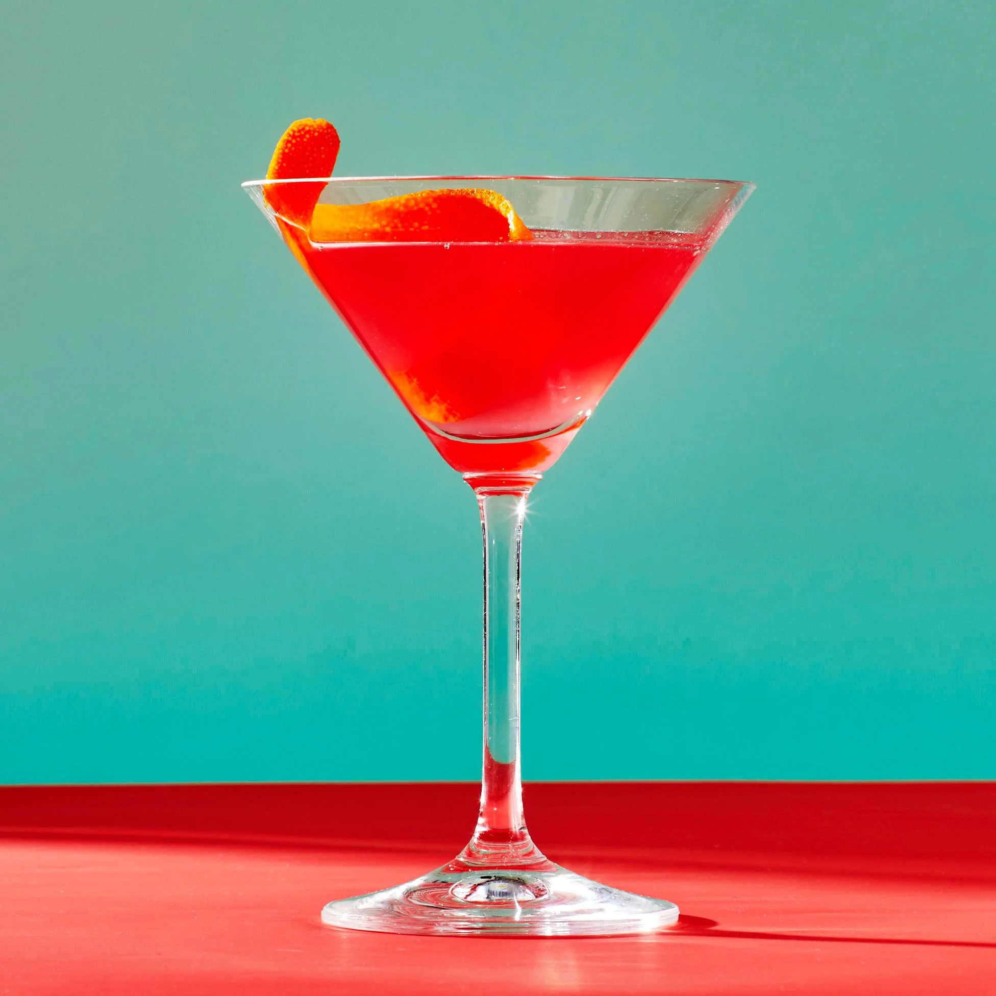 A Cosmopolitan cocktail in a martini glass garnished with an orange peel against a teal and red background