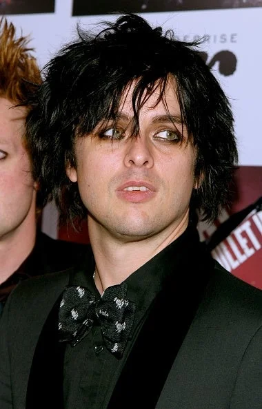 Billie Joel Armstronf with dark, tousled hair in black suit and patterned bow tie on a red carpet at an event, wearing heavy black eyeliner